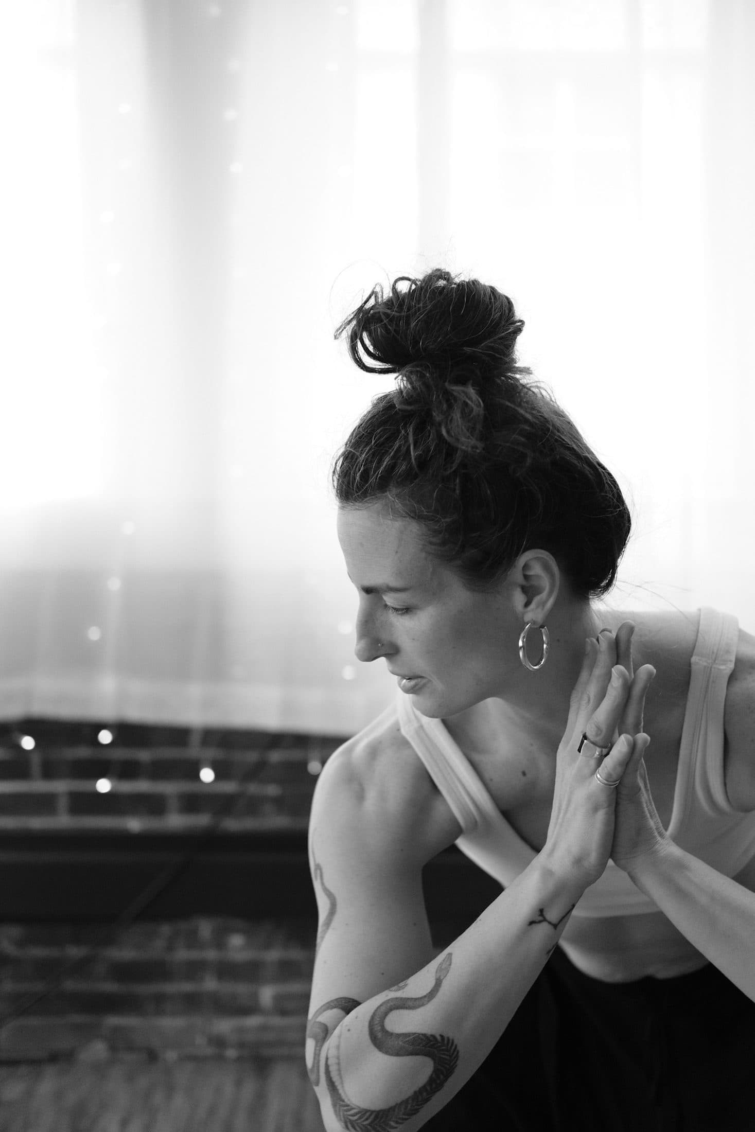 Black-and-white portrait of a practitioner in a twisting posture, hands pressed together, beside a curtained window