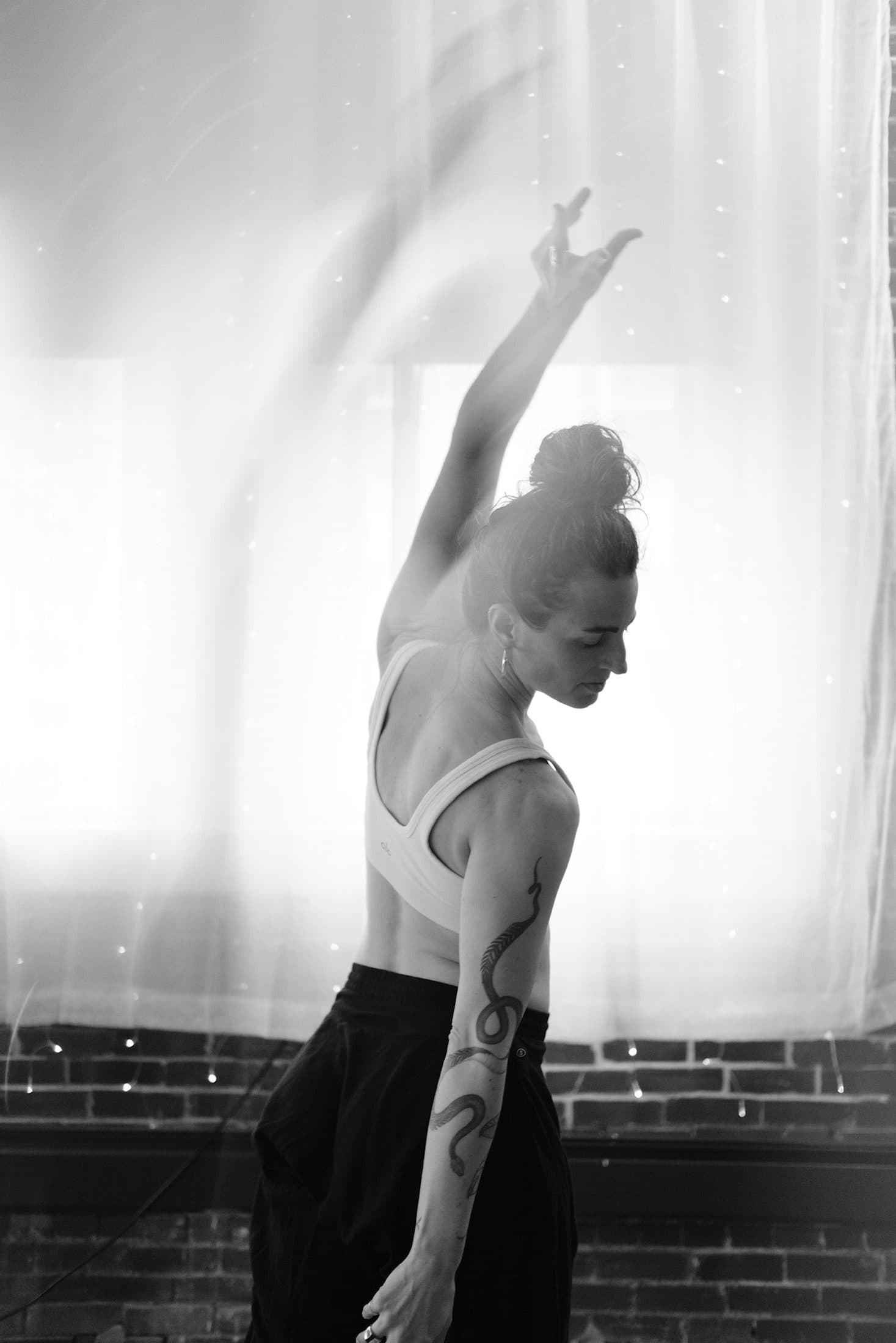 Black-and-white profile of a practitioner reaching one arm overhead against soft window light